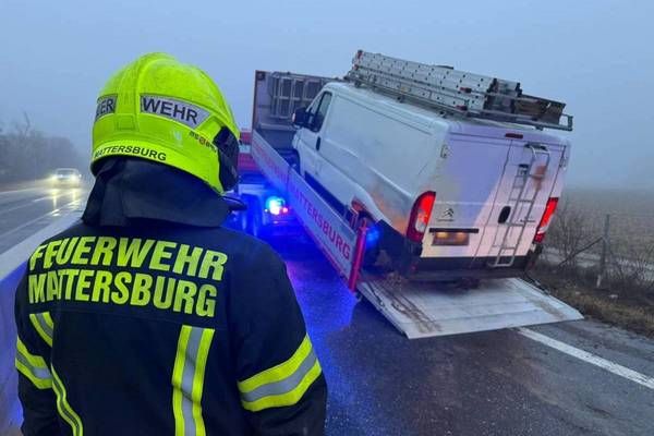 A firefighter is standing on the road, watching a white van being lifted by a tow truck. The van has a ladder on its roof and a sticker on its side.