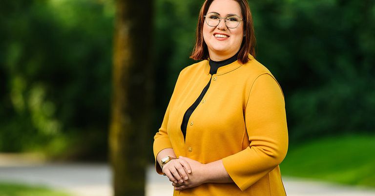 A woman with short red hair, wearing glasses, a yellow blazer, and a black top, standing with her hands clasped in front of a tree.