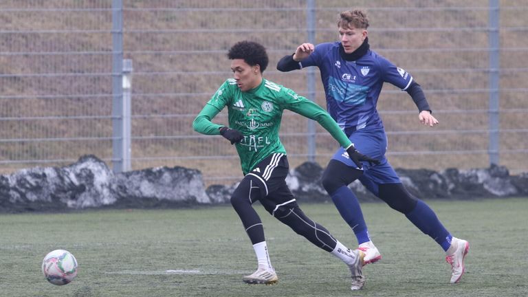 Two soccer players are running on the field. One is wearing green and the other is wearing blue. Both are wearing gloves. The field has a metal fence and a concrete barrier.