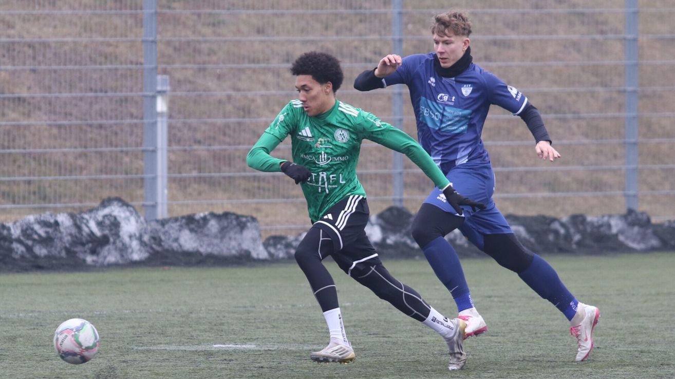 Two soccer players are running on the field. One is wearing green and the other is wearing blue. Both are wearing gloves. The field has a metal fence and a concrete barrier.