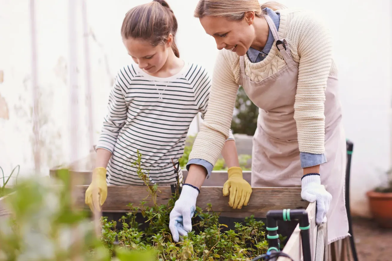 A woman and a young girl wearing gloves are working in a garden, smiling and looking down at the plants.