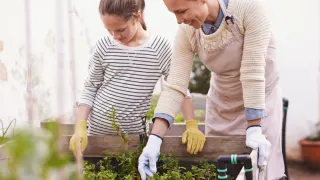 A woman and a young girl wearing gloves are working in a garden, smiling and looking down at the plants.