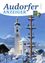 Ein Zeitschriftencover zeigt eine Kirche mit einem Uhrturm und einem verschneiten Berg im Hintergrund. Ein verzierter Pfahl mit Figuren steht in der Nähe.