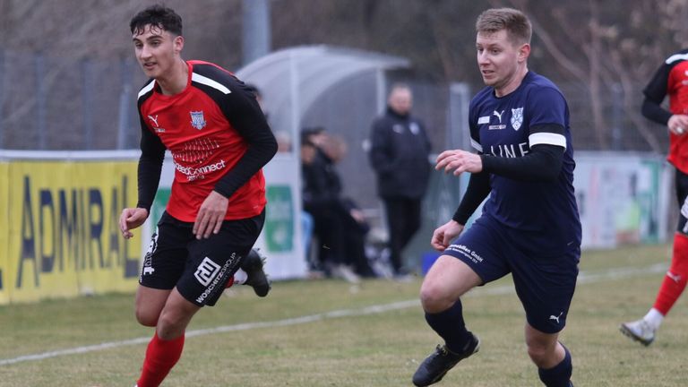 Two soccer players run towards each other, one in red and black and the other in blue. Behind them, people are sitting on the sidelines, watching the game.