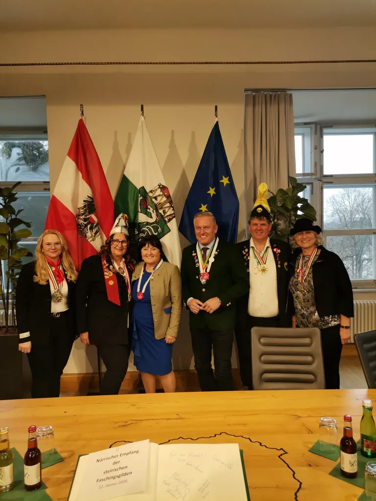 A group of individuals dressed formally stands in front of a desk. They are surrounded by flags, including the Austrian and EU flags. The room is decorated with plants and has windows with curtains.
