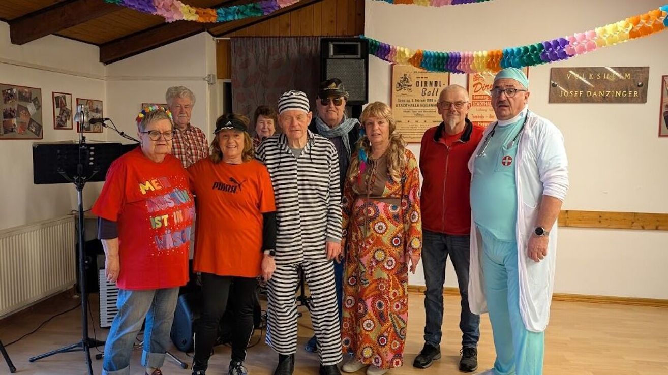 A group of adults dressed in various costumes, including a person in a nurse's outfit, are standing in a room. Colorful streamers adorn the ceiling.