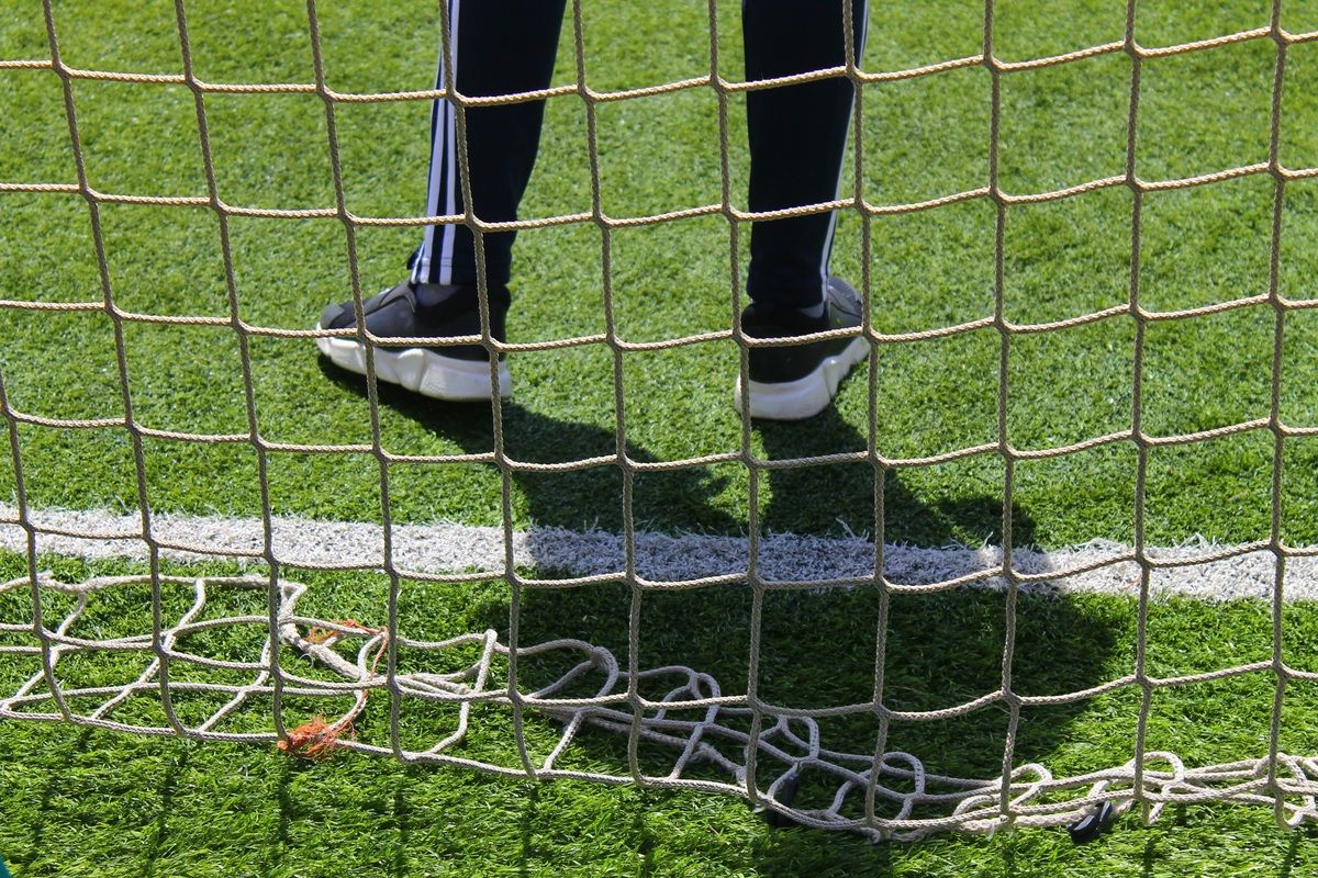 A person wearing black pants and shoes stands on a soccer field with a white net in front. The grass is green with white lines.