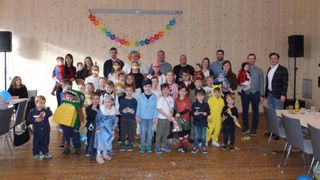 A group of children and adults wearing costumes and masks are posing for a photo. Some are holding gifts. The room has wooden walls and a colorful banner.