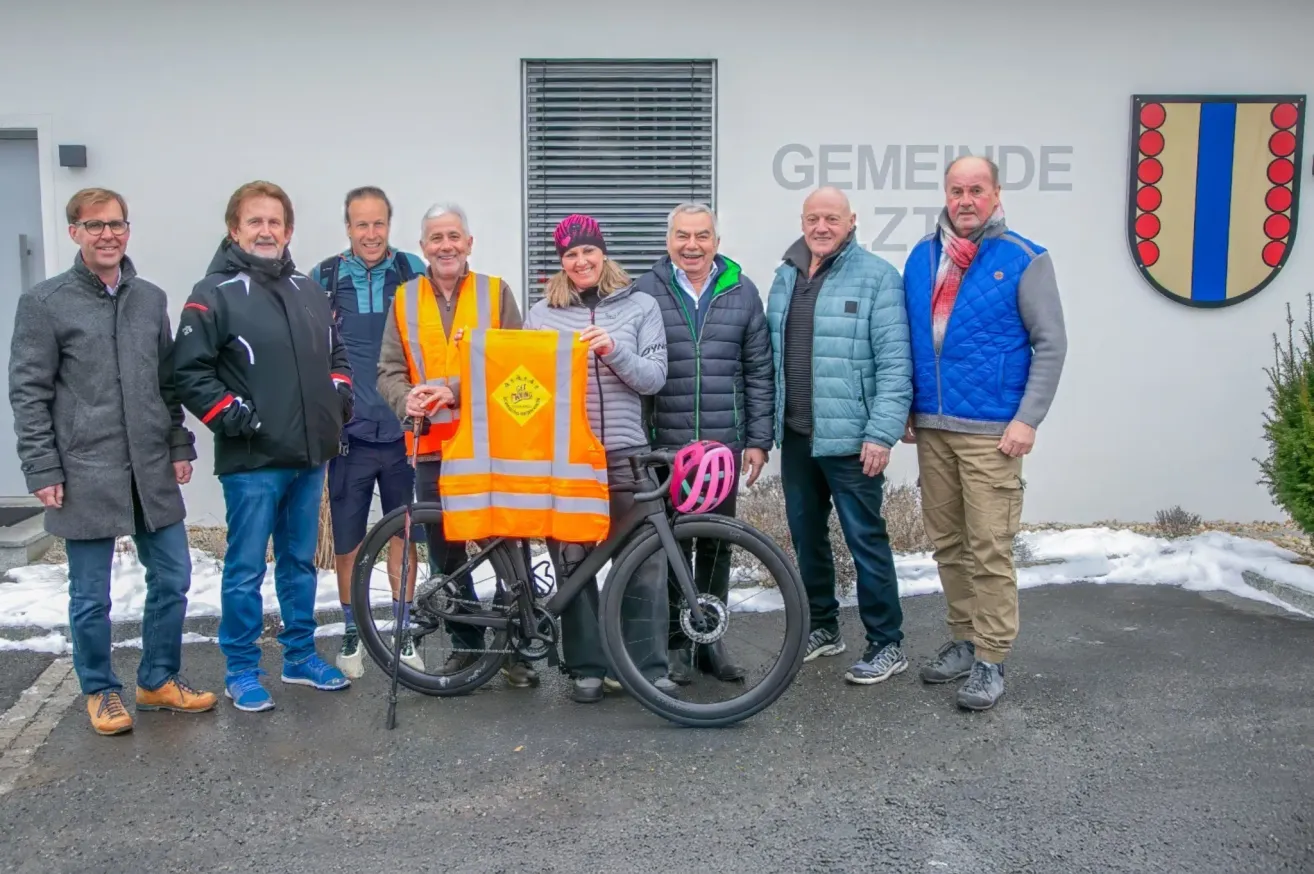Seven people are standing around a bicycle. One person is holding a reflective vest. They all appear to be smiling. The background has a sign that reads 'GEMEINDE ZIT'.