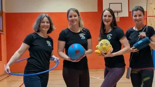 Four women in black t-shirts stand in a gymnasium, smiling. One holds a hula hoop, another a globe, and the third a stuffed toy. A basketball hoop is in the background.