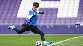 A soccer player wearing a blue jersey and black shorts practices on an empty field. The stadium seats are purple.