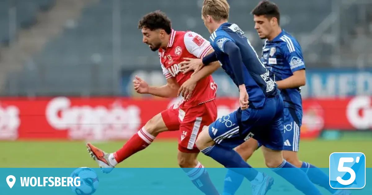Three soccer players on a field, one in red and two in blue, engage in a competitive match, with the player in red dribbling the ball and the blue players attempting to intercept.
