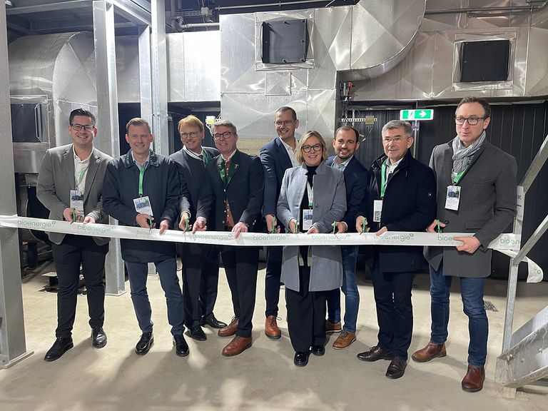 A group of people in formal attire are gathered around a ribbon, possibly for a ribbon-cutting ceremony. They are all smiling and looking at the camera. Behind them is a large metallic structure, possibly a factory.