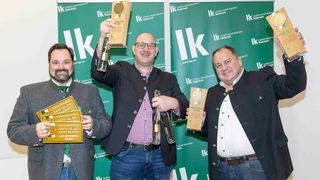 Three men stand in front of a green backdrop with an 'IK' logo. They are holding awards and bottles of alcohol. Two men hold awards with 'SafT - MOST Edelbrand 2026' text. The man in the middle holds a bottle and a wooden award.