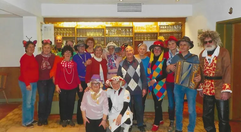 Eine Gruppe älterer Erwachsener in festlicher Kleidung, einschließlich Hüten und Halsketten, versammelt sich in einem Raum mit Regalen voller Gläser dahinter. Einige lächeln und posieren für ein Foto.