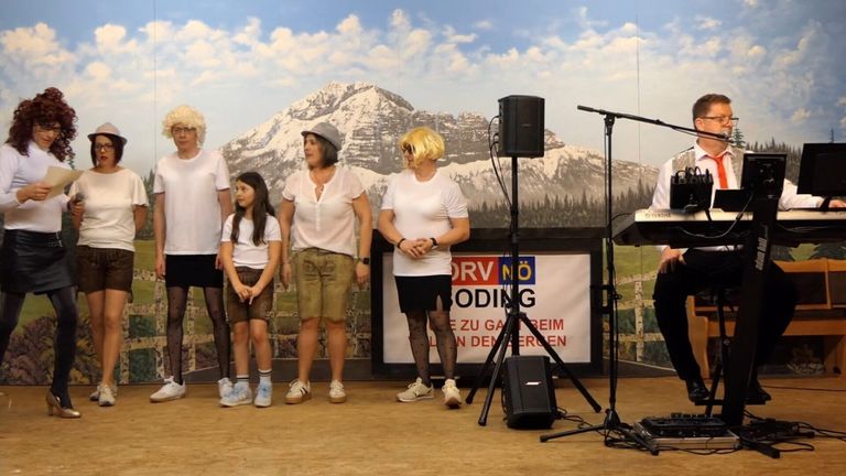 Eine Gruppe von Menschen auf einer Bühne mit einem Hintergrund von Bergen. Vier Erwachsene und ein Kind, alle in weißen Shirts, posieren für ein Foto. Musikalische Ausrüstung steht in der Nähe.