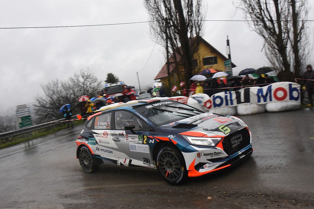 A rally car drives on a wet road, with spectators and umbrellas behind. A house with a banner is visible in the background.