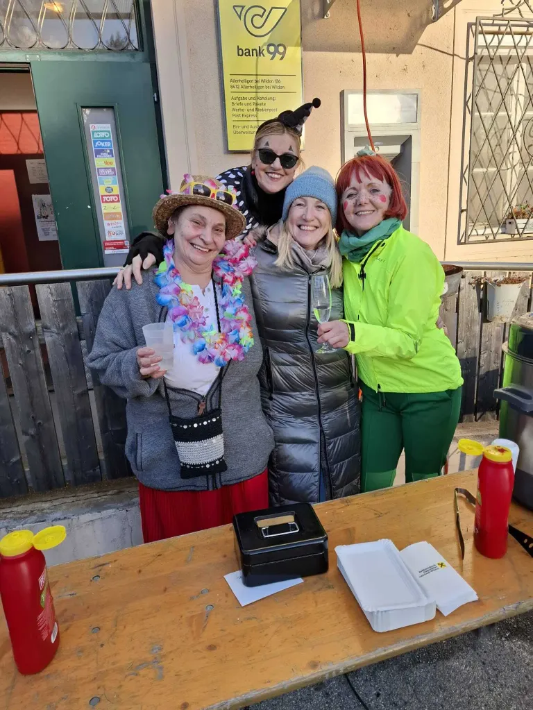 Vier Frauen, jede in festlicher Kleidung, stehen lächelnd vor einem Gebäude und halten einen Becher und ein Weinglas. Die Frau auf der linken Seite trägt einen Hut und eine Kette. Dahinter befindet sich ein Mülleimer und ein Schild an der Wand.