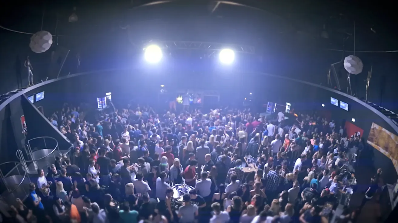 A crowded indoor venue, likely a nightclub, is illuminated by bright stage lights. The crowd is dense with people dancing and socializing, creating a lively atmosphere.