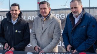 Three men stand in front of a banner. The man in the middle holds scissors, while the man on the right also holds scissors. They are all dressed formally. Behind them is a large banner with the words "Burgenland Energie". A wind turbine is visible in the background.
