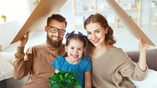 A family of three, consisting of a father, a mother, and a young girl, all smiling, standing under a tent, with the girl holding a plant.