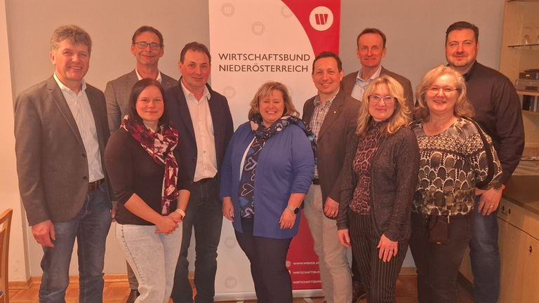 Eine Gruppe von Menschen steht lächelnd vor einem Banner. Der Schriftzug auf dem Banner lautet 'WIRTSCHAFTSBUND NIEDEROESTERREICH'. Sie sind alle in formeller Kleidung.