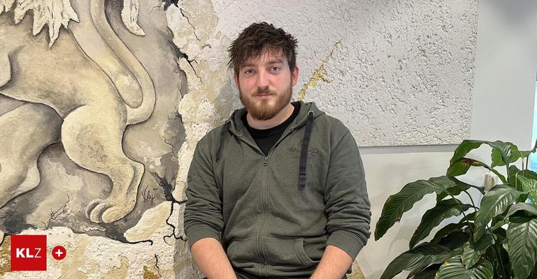 Ein Mann mit Bart in einer grünen Kapuzenjacke sitzt an einer strukturierten Wand mit einer Pflanze rechts von ihm.