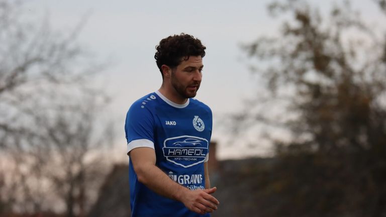 Ein Mann in einem blauen Trikot mit weißen Säumen und einem Logo auf der Brust steht auf einem Feld. Er schaut nach unten.