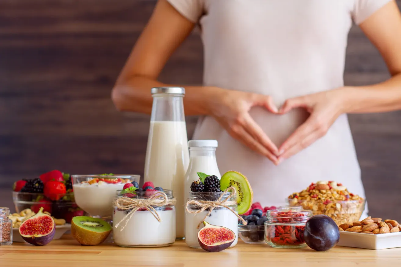A pregnant woman forms a heart shape with her hands, standing in front of a table filled with various foods and beverages, including yogurt, fruits, and milk.