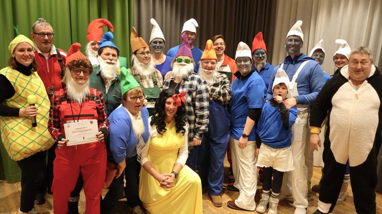A group of people in gnome costumes, with some having blue face paint, pose for a photo. A woman in a yellow dress sits in the center, smiling. The background includes a green curtain.