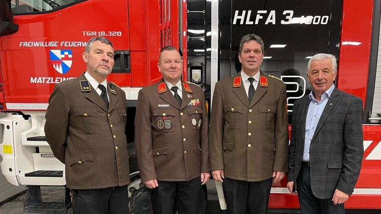 Three men in military uniforms stand in front of a red fire truck. They are all smiling and looking at the camera. The man on the right wears a tie and has medals on his chest.