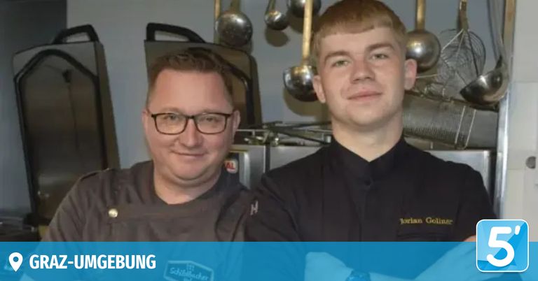 Zwei Männer in Kochuniformen stehen in einer Küche, einer mit Brille, beide lächeln. Küchengeräte und Utensilien sind im Hintergrund zu sehen.