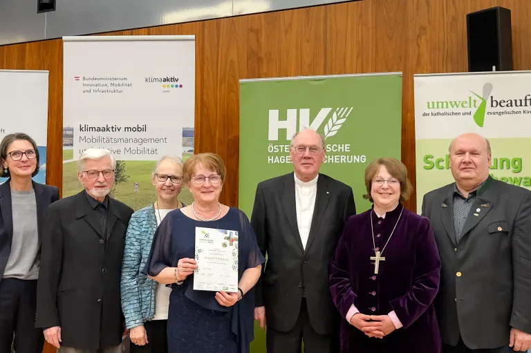 Fünf Personen stehen vor Bannern. Eine Frau hält eine Urkunde, während die anderen eine Brille und Halsketten tragen. Sie stehen hinter einem Banner mit den Worten 'Hauptpreis'.