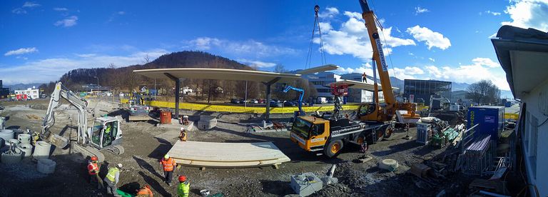 Eine Baustelle mit Arbeitern in Schutzausrüstung. Ein Kran hebt eine große Metallstruktur. Dahinter trennt eine gelbe Barriere die Baustelle. Im Hintergrund sind Berge und ein blauer Himmel mit Wolken.