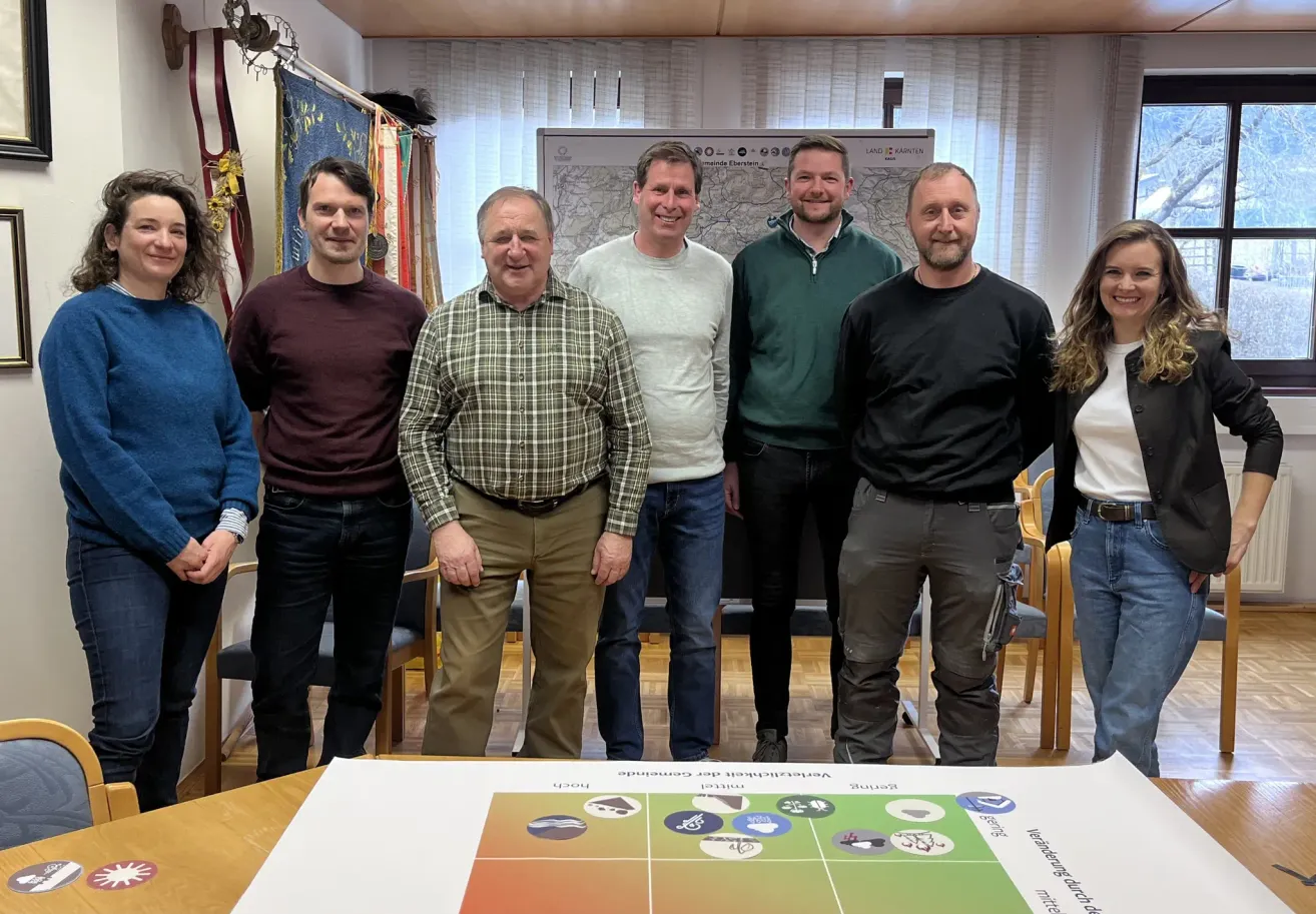 Sechs Männer und eine Frau stehen in einem Raum und lächeln für ein Foto. Hinter ihnen sind eine Karte und einige Flaggen zu sehen. Sie sind alle lässig gekleidet.