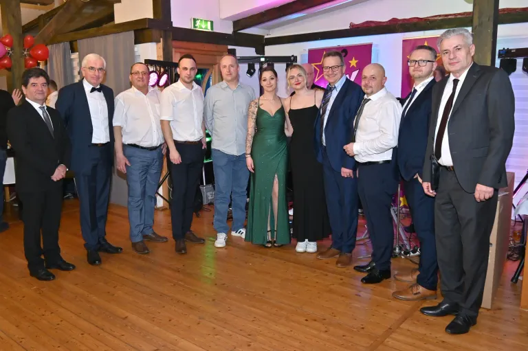 Eine Gruppe formell gekleideter Männer und Frauen steht auf einem Holzboden, wahrscheinlich bei einer Veranstaltung. Zwei Frauen stehen in der Mitte, eine trägt ein grünes und die andere ein schwarzes Kleid. Hinter ihnen hängen Flaggen.