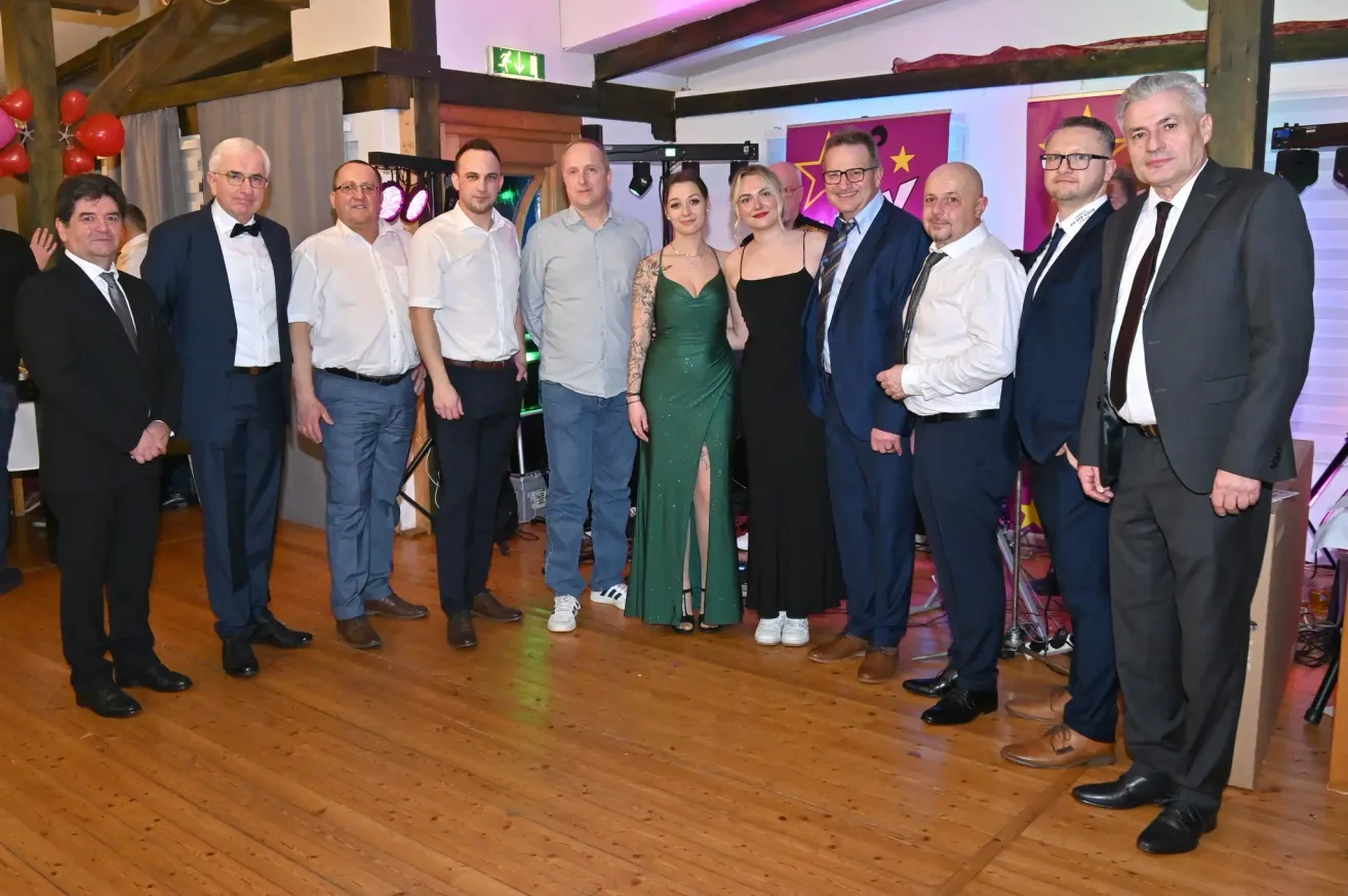 Eine Gruppe formell gekleideter Männer und Frauen steht auf einem Holzboden, wahrscheinlich bei einer Veranstaltung. Zwei Frauen stehen in der Mitte, eine trägt ein grünes und die andere ein schwarzes Kleid. Hinter ihnen hängen Flaggen.