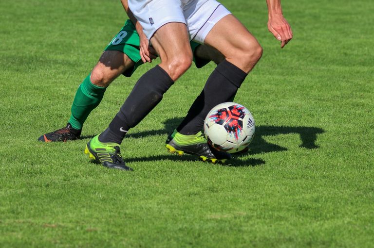 Two soccer players compete on a field, one wearing green and the other in white. The player in white kicks the ball.