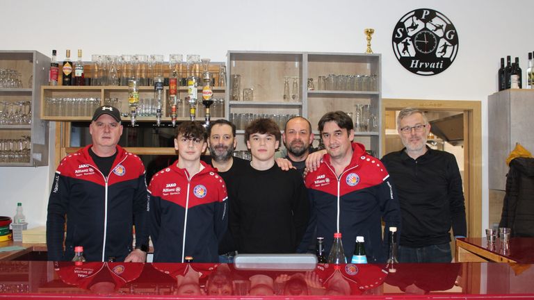 Sechs Männer, darunter zwei junge Athleten in roten und schwarzen Jacken, posieren in einer Bar. Die Umgebung besteht aus Regalen mit Gläsern, einem Pokal und einer Uhr. Flaschen und Gläser stehen auf der Theke.