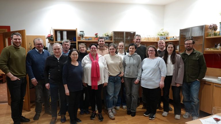 Eine Gruppe von Menschen, sowohl Männer als auch Frauen, stehen zusammen in einem Raum mit Holzboden. Sie scheinen für ein Foto zu posieren. Hinter ihnen befinden sich Regale mit verschiedenen ausgestellten Gegenständen.