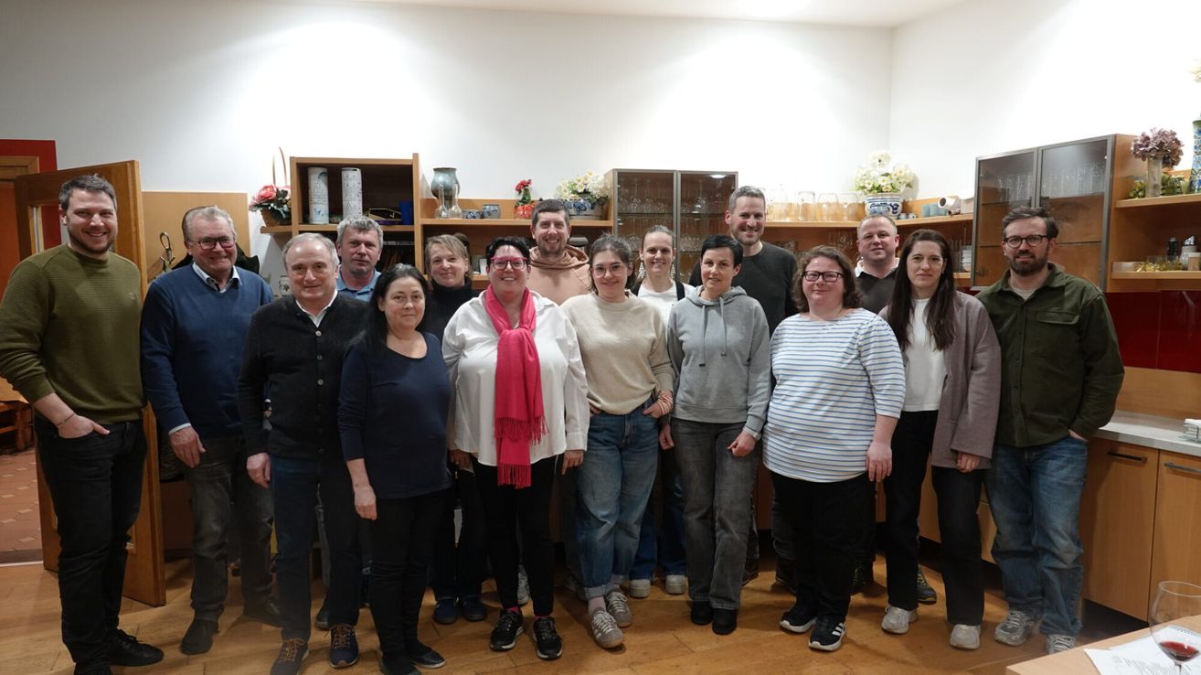 Eine Gruppe von Menschen, sowohl Männer als auch Frauen, stehen zusammen in einem Raum mit Holzboden. Sie scheinen für ein Foto zu posieren. Hinter ihnen befinden sich Regale mit verschiedenen ausgestellten Gegenständen.