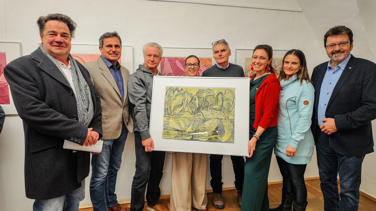 A group of people stand around a framed artwork, smiling for a photo. They are in a room with wooden floors and several paintings on the wall.