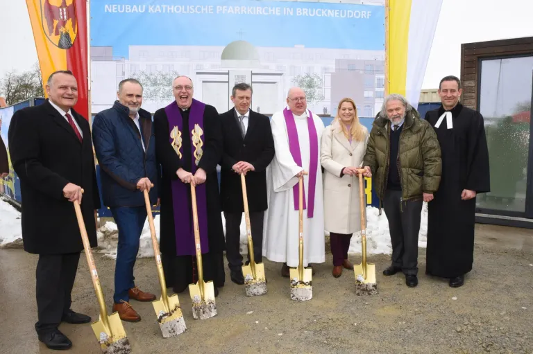 A group of people in formal attire hold shovels for a groundbreaking ceremony, standing in front of a banner for the new Catholic parish church in Bruckneudorf.