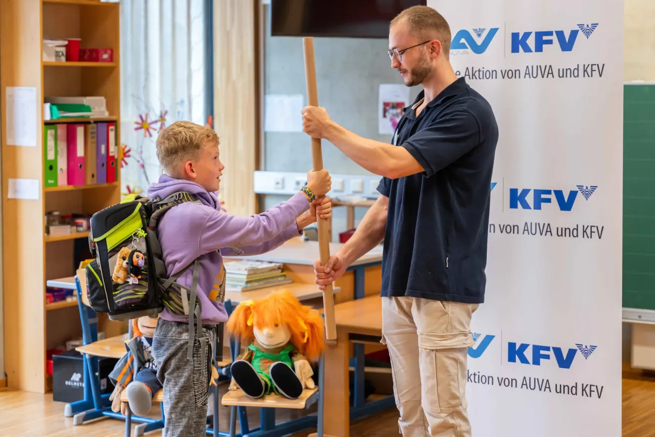 A man in glasses and a black shirt holds a wooden stick while a boy in a purple shirt holds the other end. The boy has a backpack and there are stuffed toys on a chair.