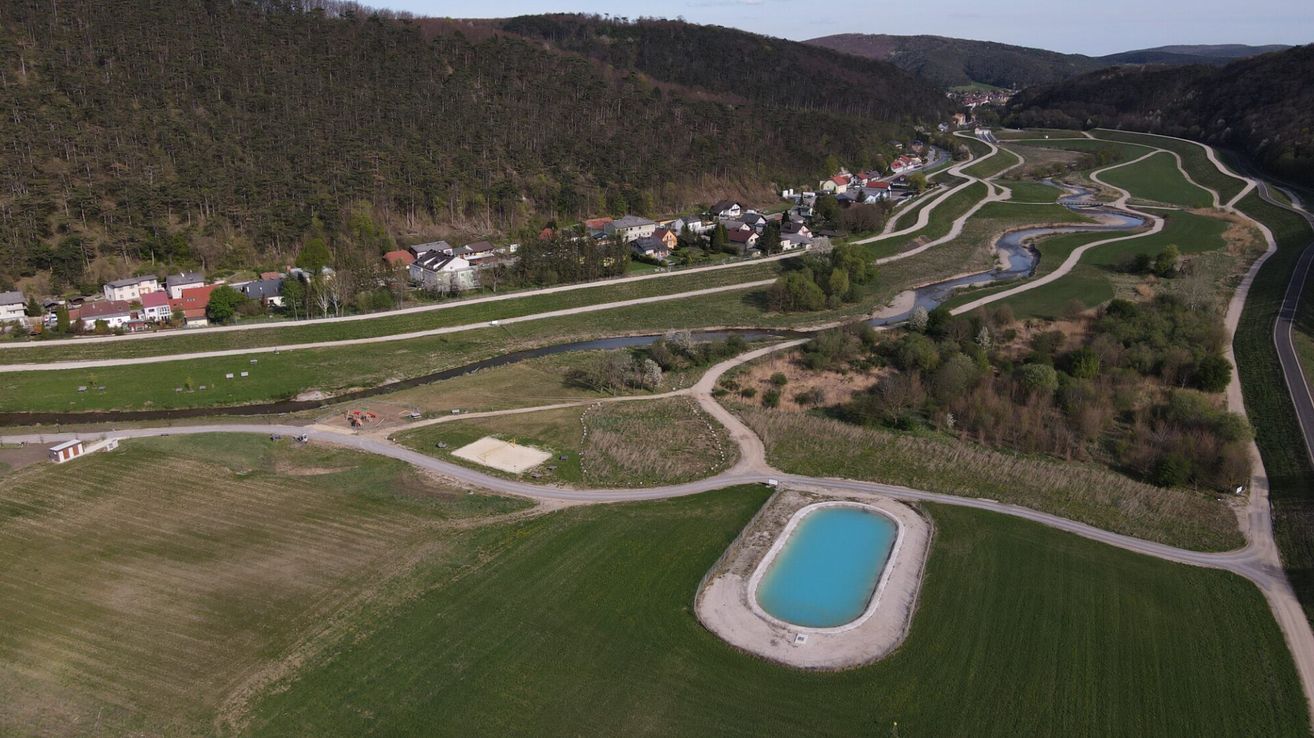 Luftaufnahme einer ländlichen Gegend mit einem kleinen Dorf, einem mäandernden Fluss und einem neu errichteten Pool mit klarem blauem Wasser in einem grünen Feld.
