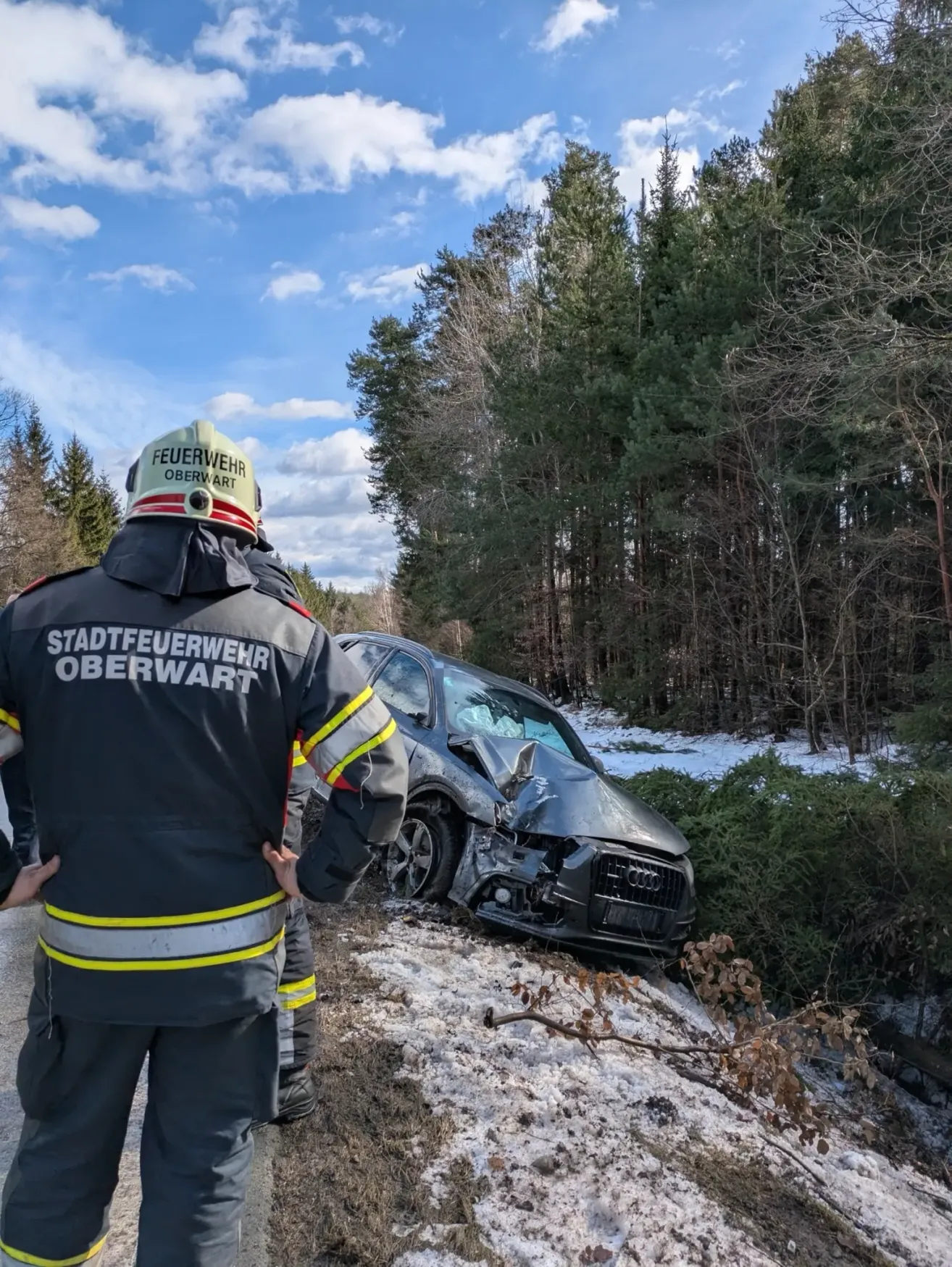 Zwei Feuerwehrleute stehen neben einem verunglückten Auto in einem verschneiten Gebiet. Einer trägt einen Helm mit dem Wort 'Feuerwehr'.