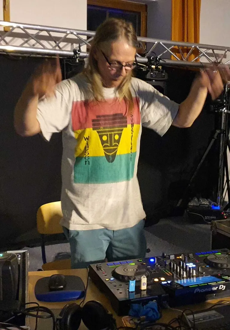 A man with glasses and long hair stands behind a DJ console, wearing a white t-shirt with a colorful design. A yellow chair is behind him.