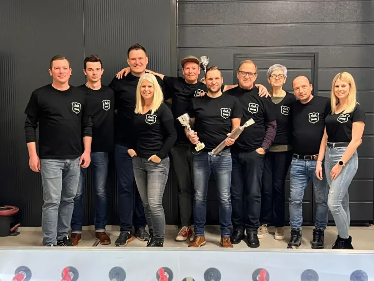 Eine Gruppe von Menschen in gleichen schwarzen T-Shirts mit weißen Logos posiert für ein Foto. Einige halten Trophäen, und sie stehen auf einer Bühne mit einer grauen Wand im Hintergrund.