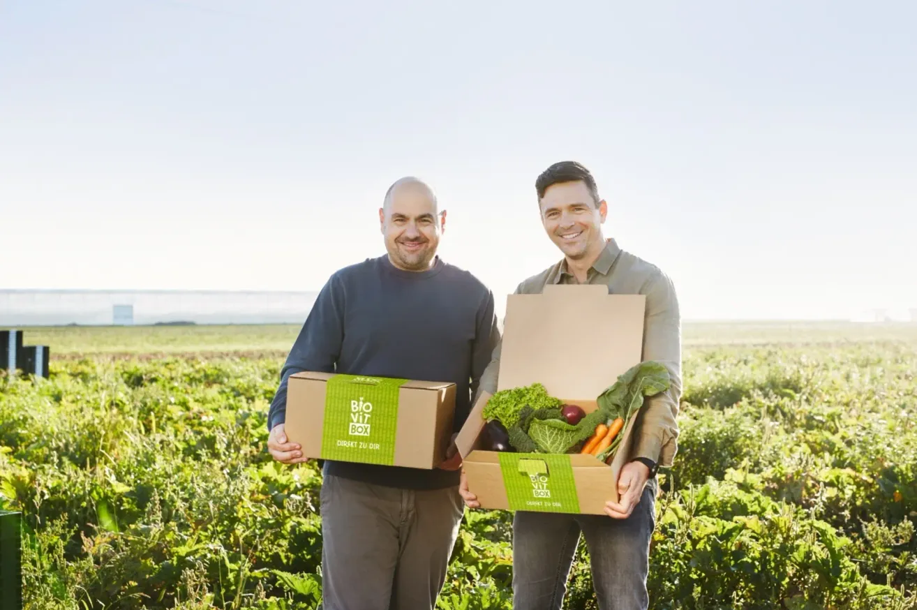 Zwei Männer auf einem Feld halten eine Box mit der Aufschrift 'BIO VOiT BOX' und eine weitere Box mit Gemüse.