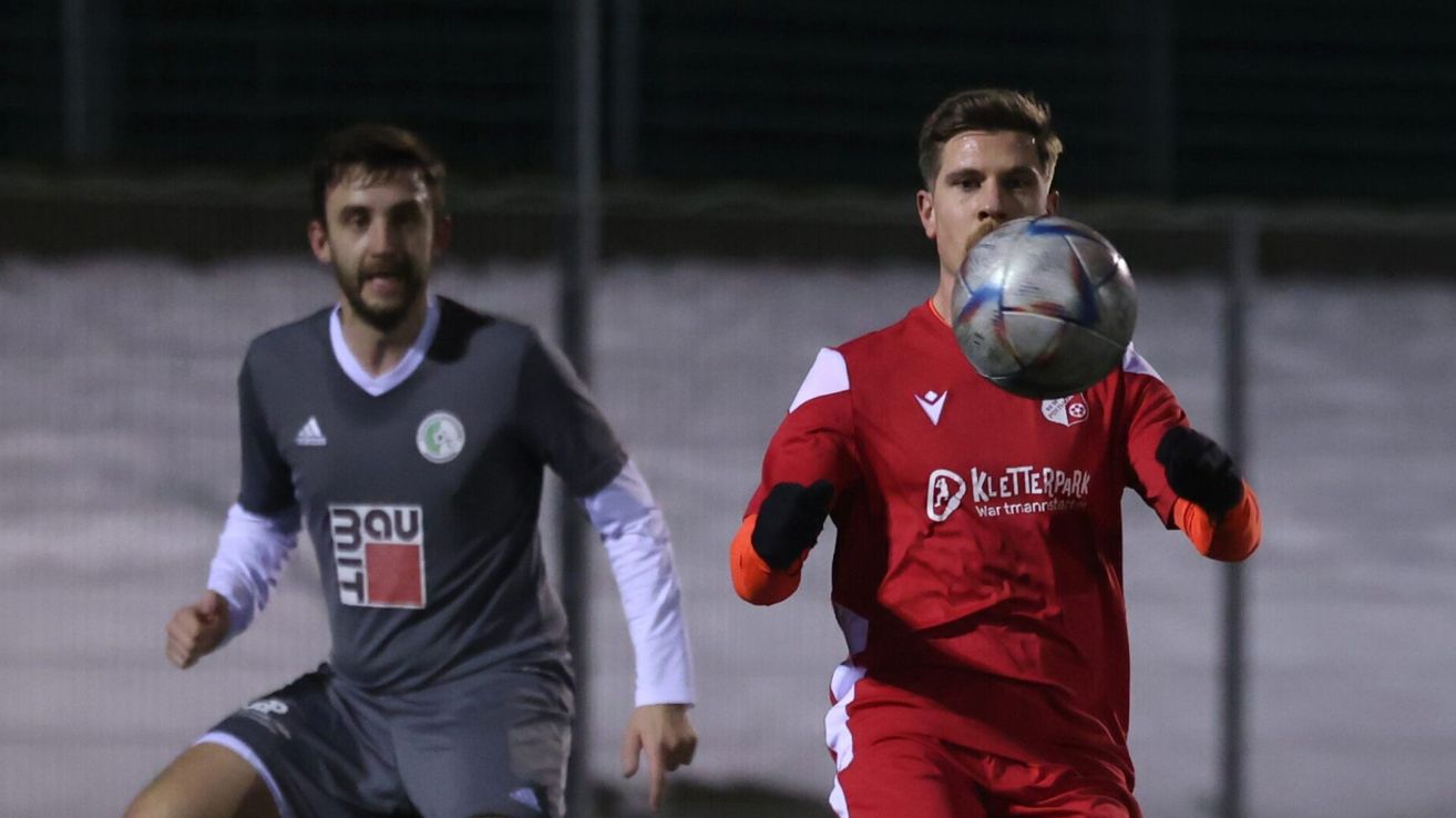 Two men play soccer. One wears a gray shirt with a logo, the other a red shirt with a logo. One holds a soccer ball.